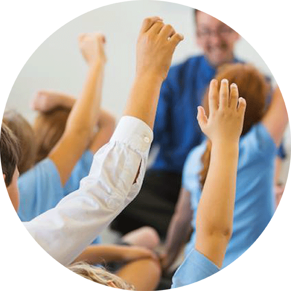 Class of school children raising their hands before a young male Early Careers Teacher