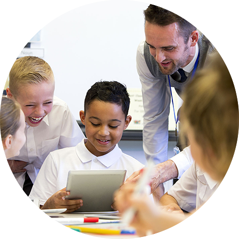 Male teacher leaning over young student using tablet device while other students observe